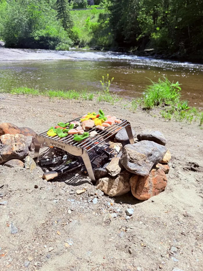 OakStoke Open Fire Grill cooking chicken and vegetables over a campfire on a riverbank, 18x12 stainless steel open fire cooking grill