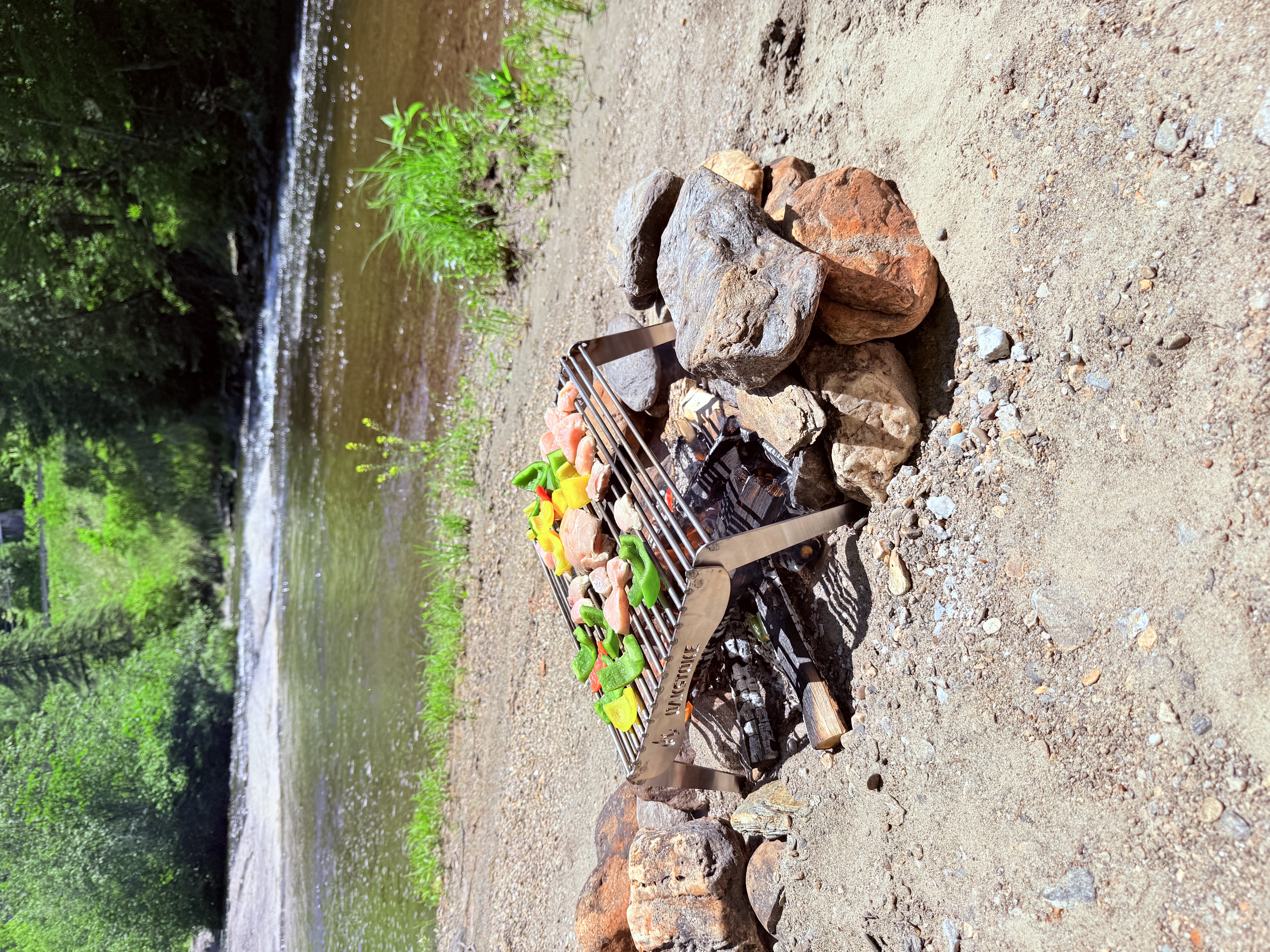 OakStoke Open Fire Grill cooking chicken and vegetables over a campfire on a riverbank, 18x12 stainless steel