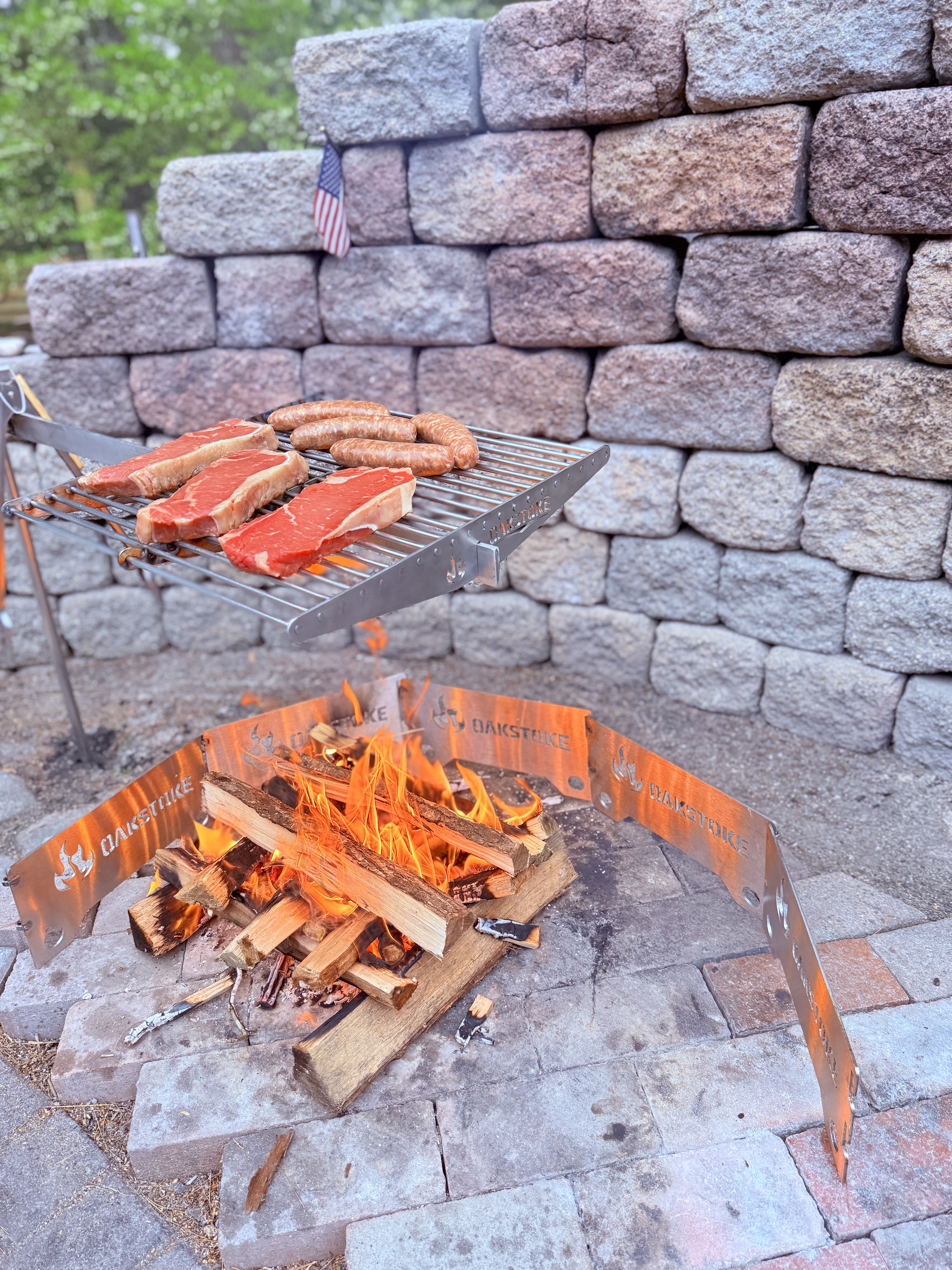 OakStoke Campfire Swivel Arm with steaks and sausages cooking over open fire, stainless steel campfire grill stand in use
